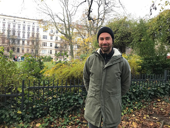 A man in a black beanie and olive green parka smiling in a park with buildings in the background.