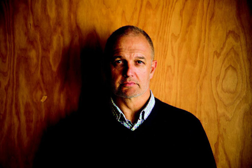 A man in a dark sweater stands against a warm-toned wood-paneled wall in dramatic lighting.