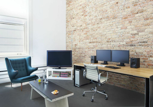 A workspace featuring a wooden desk with dual monitors, an office chair, and a teal armchair near a brick wall.