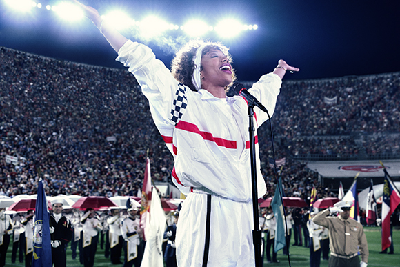 A woman in a white jumpsuit sings with arms raised before a large crowd in a stadium at night.