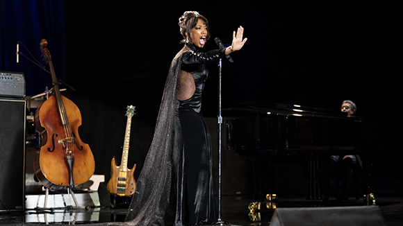 A woman in a long black gown sings into a microphone on a dark stage next to a double bass and piano.