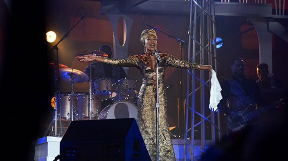A singer in a gold gown performs on stage with a drummer and bassist behind her.