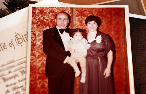 A man in a tuxedo and a woman in a dark dress hold a small child in front of a red patterned backdrop.