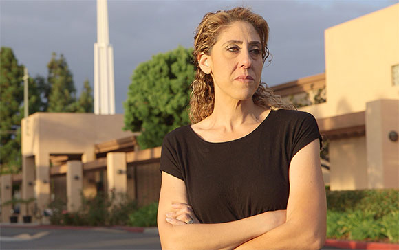A woman with curly hair and a black t-shirt stands with her arms crossed in front of trees and buildings.