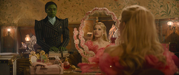 A green-skinned woman in a dark suit stands behind a blonde woman reflected in a pink ornate mirror.