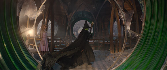 A person in a black cloak and pointed hat stands in a large, circular wooden room seen through broken glass.