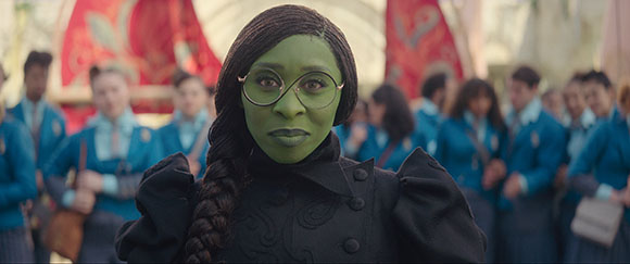 A green-skinned woman with dark braided hair and round glasses stands before a crowd in blue uniforms.