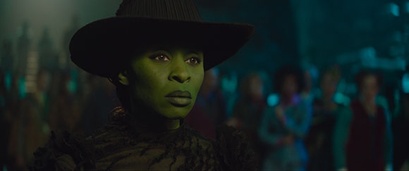 A woman with green skin and dark eyes wearing a large dark hat, looking forward with a serious expression.