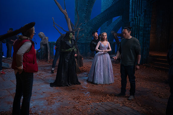 Actors in costume and crew members stand on a set covered in autumn leaves under blue lighting.