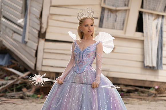 A woman in a shimmering lavender gown and silver crown holds a star-topped wand outdoors.