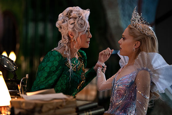 A woman in a green gown and silver hair leans toward a blonde woman in a shimmering lavender dress.