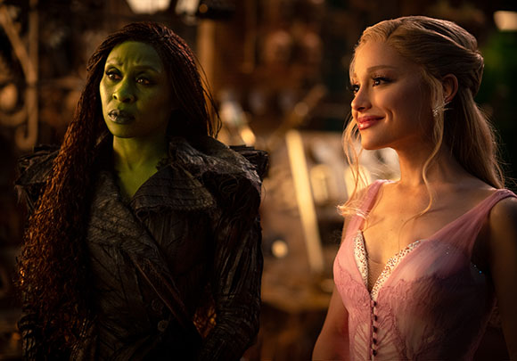 A green-skinned woman in dark clothing looks skeptically at a blonde woman in a pink dress.