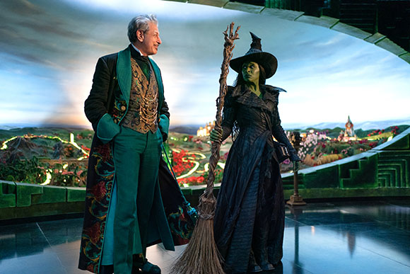 A man in an ornate green suit smiles at a green-skinned woman wearing a black witch hat and holding a broom.