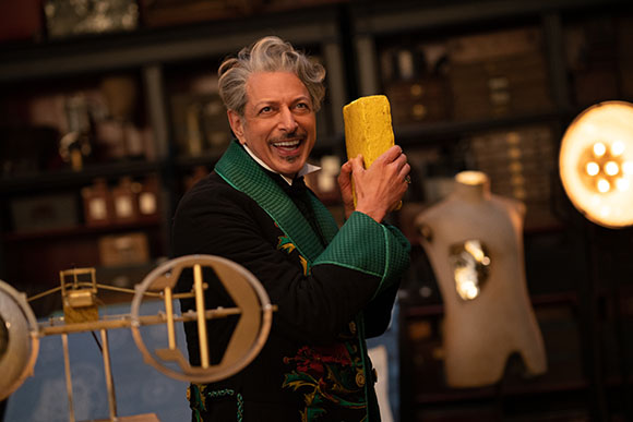 A man with grey hair smiles while holding a yellow block in a workshop filled with vintage items.