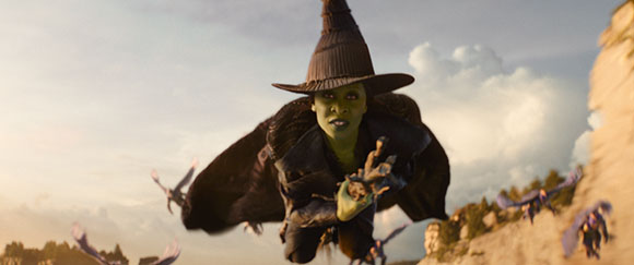 A green-skinned woman wearing a black pointed hat and cloak flies through a cloudy sky.