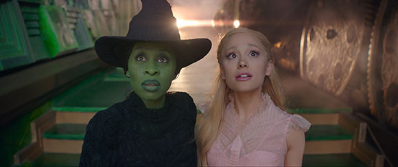 A green-skinned woman in a black witch hat and a blonde woman in a pink dress look upward in awe.