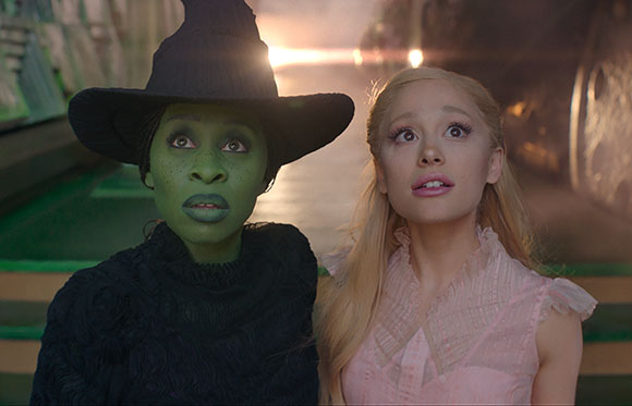 A woman in green skin and a black witch hat stands next to a blonde woman in a pink ruffled top.