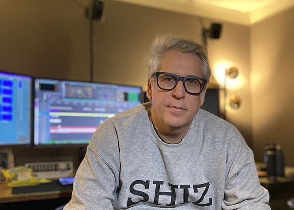 A man with grey hair and glasses wearing a grey sweatshirt in a studio setting.