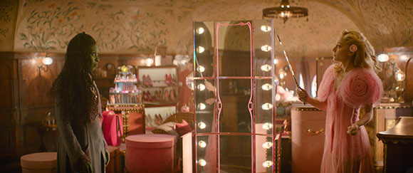 A woman in a pink dress holds a wand toward a woman in a grey outfit in a vintage dressing room.