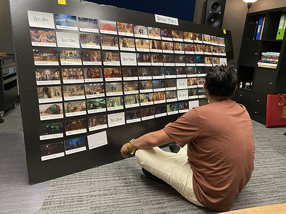 A person in a brown shirt sits on the floor, viewing a large black board covered in rows of film storyboard images.
