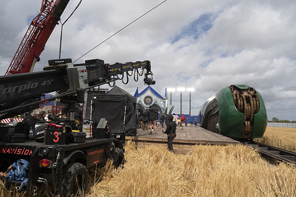 A large camera crane and film equipment stand on a wooden platform in a field with crew members nearby.