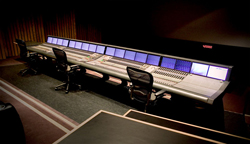 A long, professional audio mixing console with multiple screens and three black office chairs in a dark studio.