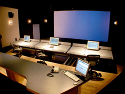 Control room with several computer desks, monitors, and a large blue projection screen in a dark room.