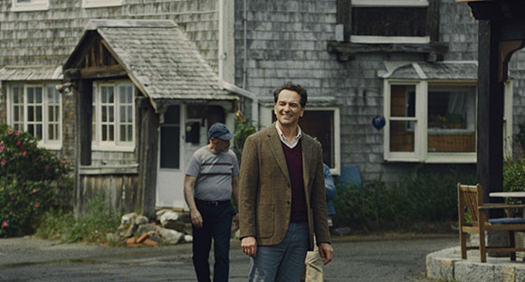 A man in a brown blazer and maroon sweater walks toward the camera on a street near weathered wooden houses.
