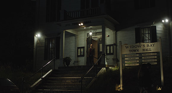 A person stands in the lit doorway of a white wooden building labeled Widow's Bay Town Hall at night.