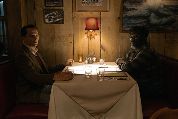 Two men sit opposite each other at a white-clothed table in a dimly lit restaurant.