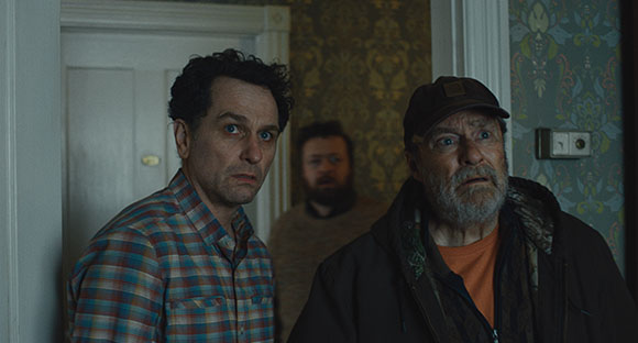 Three men with worried expressions stand in a dimly lit room near a doorway.