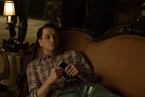 A man in a plaid shirt reclines on an ornate brown sofa in a dimly lit room.