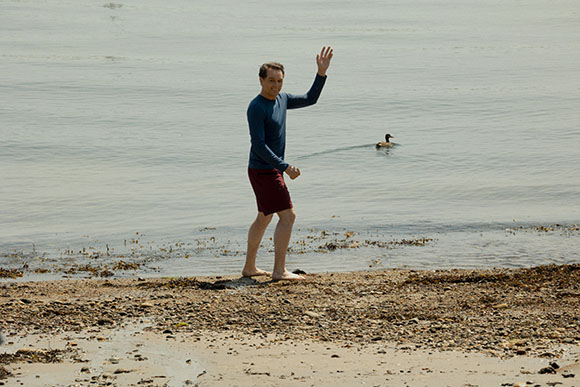 A man in a blue long-sleeve shirt and maroon shorts waves while walking along a shoreline.