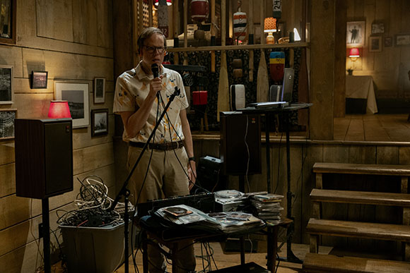 A man in a patterned shirt speaks into a microphone behind a table of records and audio equipment.