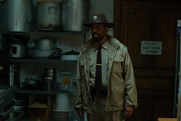 A man in a tan park ranger uniform and wide-brimmed hat stands in a dimly lit kitchen.