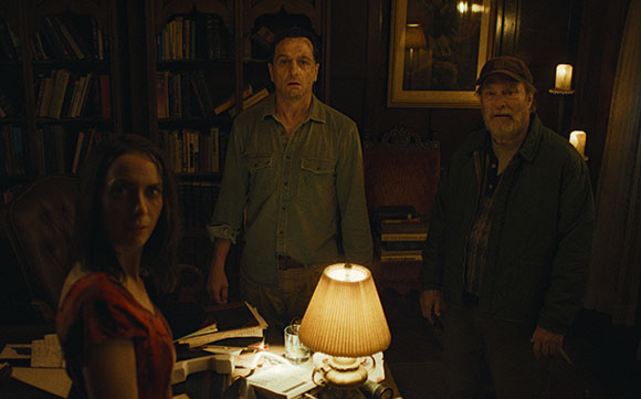 A woman and two men stand in a dark library, looking toward the camera with expressions of concern.