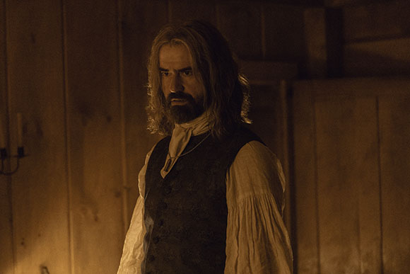 A man with long wavy hair and a beard wearing a dark vest over a white poet shirt in dim lighting.