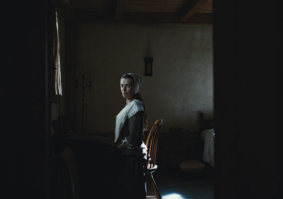 A woman wearing a white bonnet and period dress stands in a dark, rustic room.