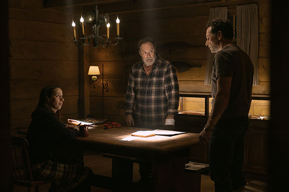 A woman sits at a table while a man stands opposite her and another man stands in the center of a cabin.