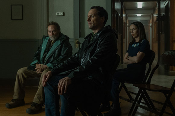 A man in a leather jacket sits in the foreground, flanked by an older man and a woman in a dimly lit hallway.