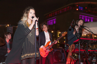 A female singer holds a microphone while a guitarist and keyboardist perform on a stage at night.