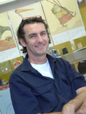 A smiling man with dark curly hair wearing a blue button-down shirt over a white t-shirt.