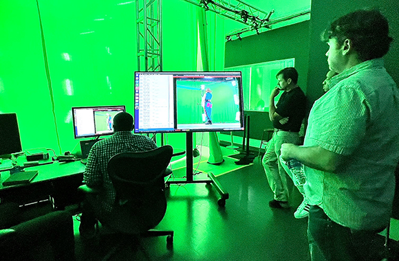 Film crew monitors a green screen studio setup with large screens displaying a person in front of a green backdrop.