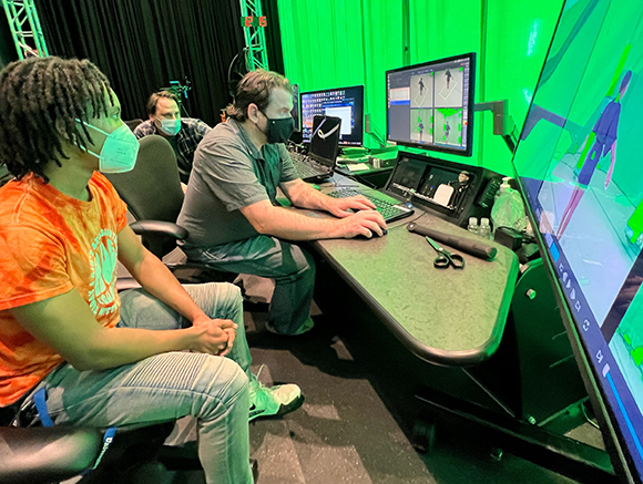 Three people in a studio with a green screen, monitors, and computer equipment.