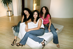Three women sitting together on a large white ottoman in a brightly lit room.