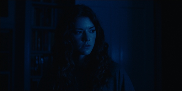 A woman with long wavy hair looks pensively to the side in a dark, blue-toned room.