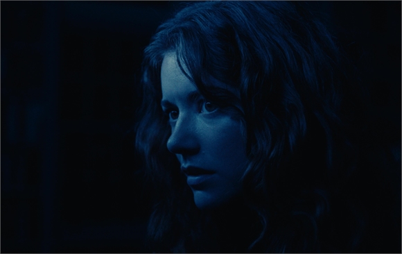 Close-up of a woman with wavy hair looking off to the side in dim, blue lighting.