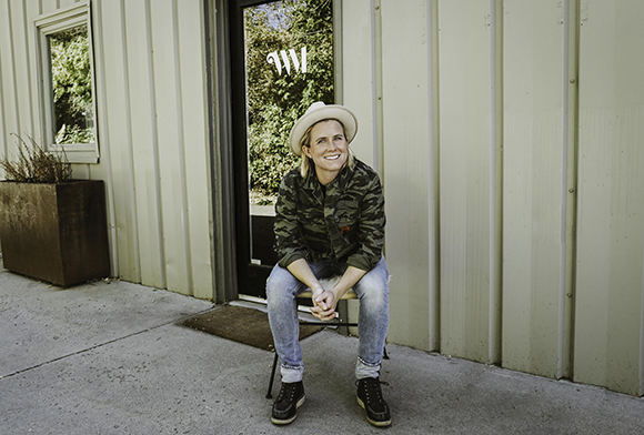 A smiling woman wearing a wide-brimmed hat and camouflage jacket sits on a chair in front of a building.