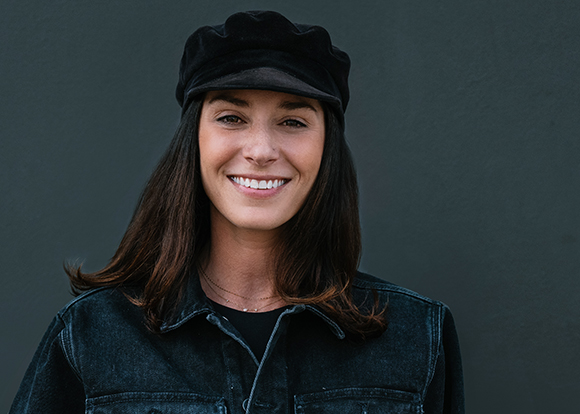 A smiling woman with long brown hair wearing a black velvet cap and a dark denim jacket against a dark wall.