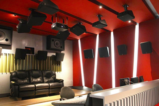 A sound studio with red walls, black leather seating, and numerous black speakers mounted on the ceiling and walls.